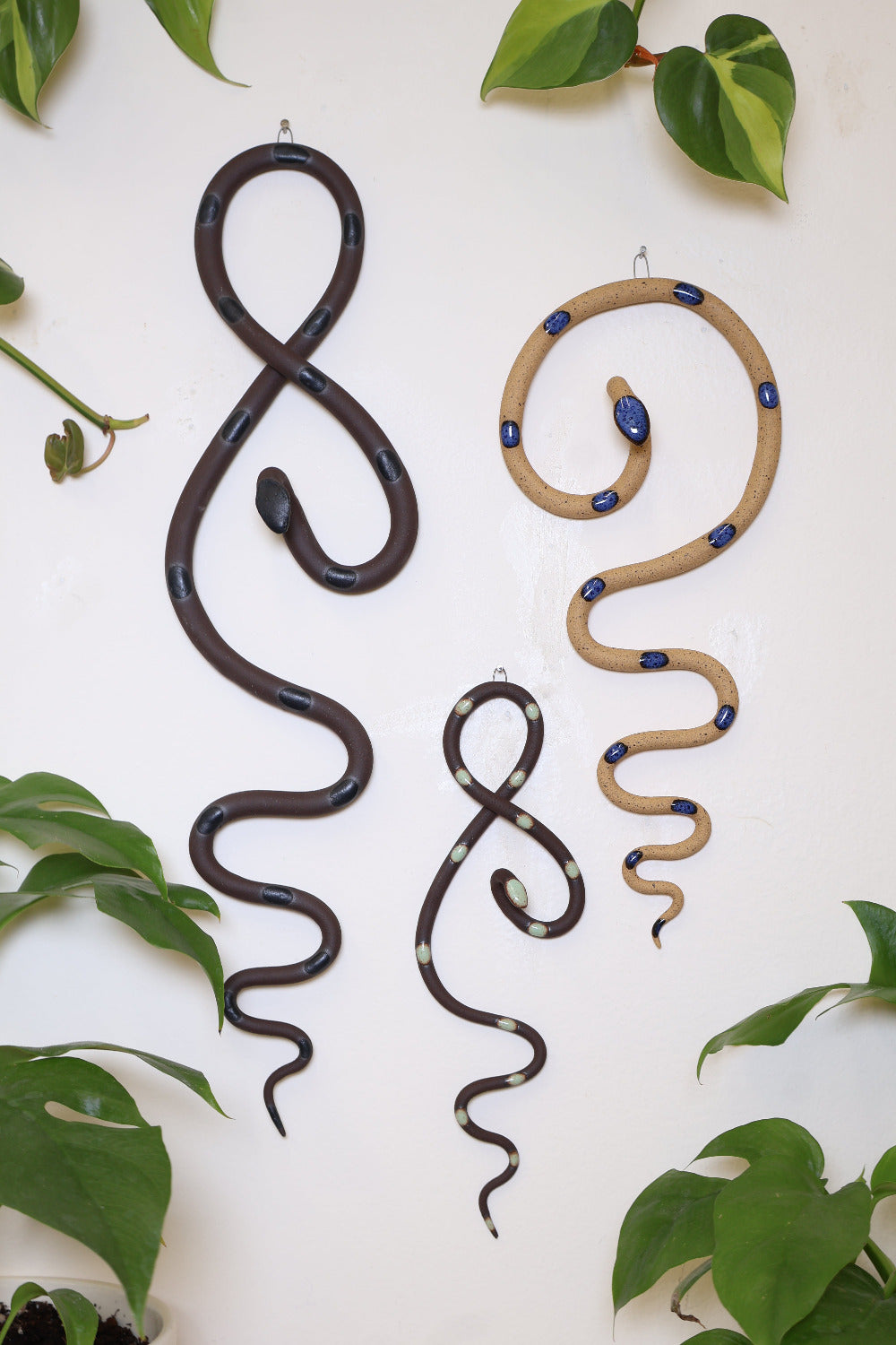 Marsh curated set of three wall ceramic snakes. From left to right hanging on a wall is a Saul wall snake (dark chocolate clay and black glaze), a mini Stevie wall snake (dark chocolate clay and turquoise glaze), and a medium Selena wall snake (speckled tan clay and blue glaze).