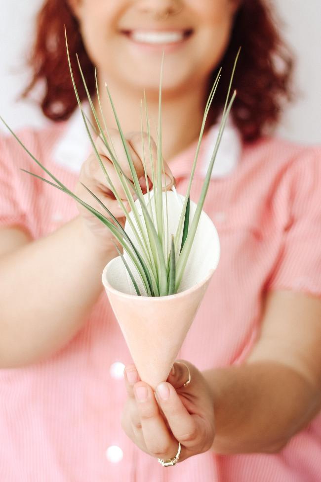 Contemporary ivory clay formed cone with light pink glaze styled with a spikey green air plant. The cone is being held by a young smiling woman showing the faux leather loop at the top for hanging the cone.  