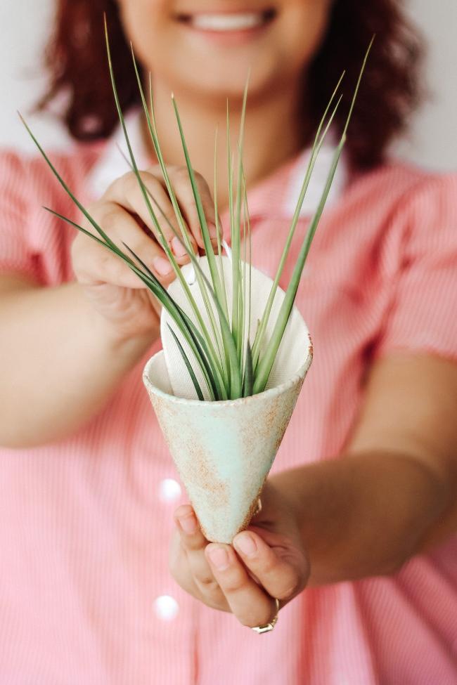 Contemporary ivory clay formed cone with light turquoise glaze styled with a spikey green air plant. The cone is being held by a young smiling woman showing the faux leather loop at the top for hanging the cone.  