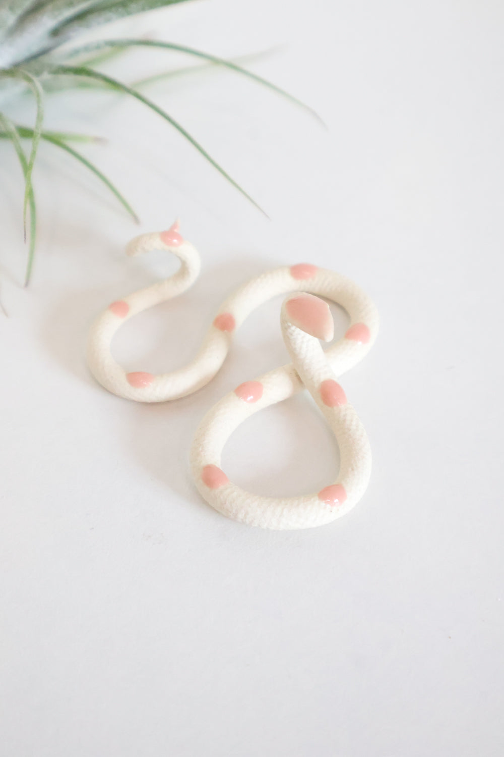 Itty bitty organic s-shaped pottery snake sculpture in the colorway Sofie, ivory white clay body with a light pink glazed head and evenly spaced glazed dots along its body.