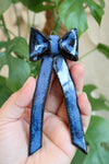 A hand holding a glossy ceramic ornament in the shape of a bow. The ornament is a deep blue with black edges, featuring a speckled texture. A leafy green plant is visible in the softly blurred background