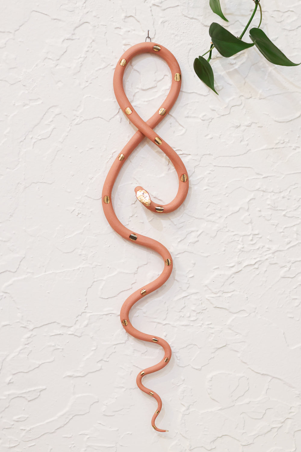 Desert sand clay vertical wall snake sculpture with gold glazed  head and dots. The top of the snake crosses like a figure 8 and the tail hangs down in an organic s curve. It hangs vertically from a small metal loop on a white wall with vining pothos. 