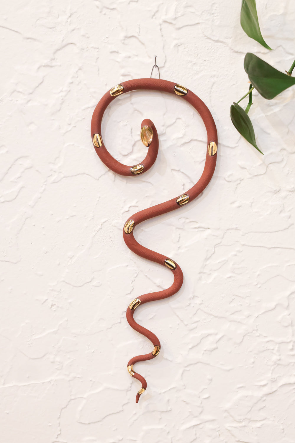 Red terracotta coiled pottery wall snake with gold glazed head and dots. The snake is shaped loosely like a question mark with the tail hanging down in an organic s curve. 