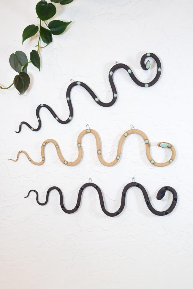 Three horizontal ceramic wavy snakes hanging on a white wall. The top snake is white clay with light turquoise glazed dots. The middle snake is a dark brown clay with light turquoise glazed dots. The bottom snake is tan clay with light turquoise glazed head and dots. 