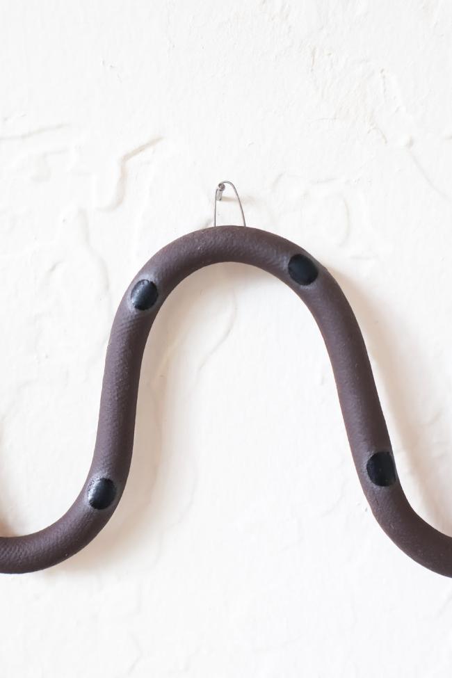 A close up of one of the ceramic snake curves showing the Saul colorway, dark brown clay base and black dots. It hangs on a nail from a small metal loop.