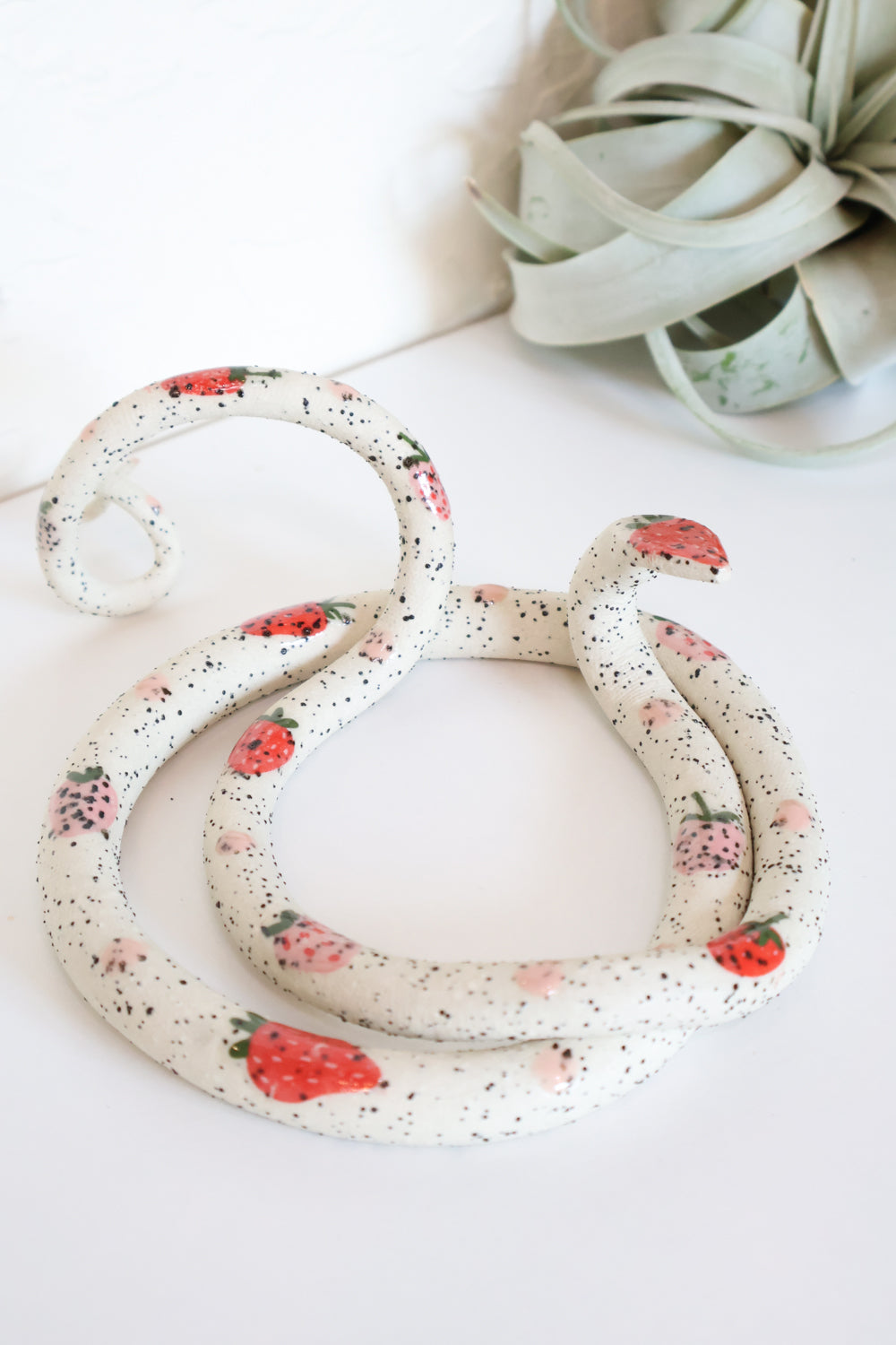 Ivory clay snake with black speckles and pink and red strawberries. The snake is coiled in a circle with it's head raised and tail in a spiral