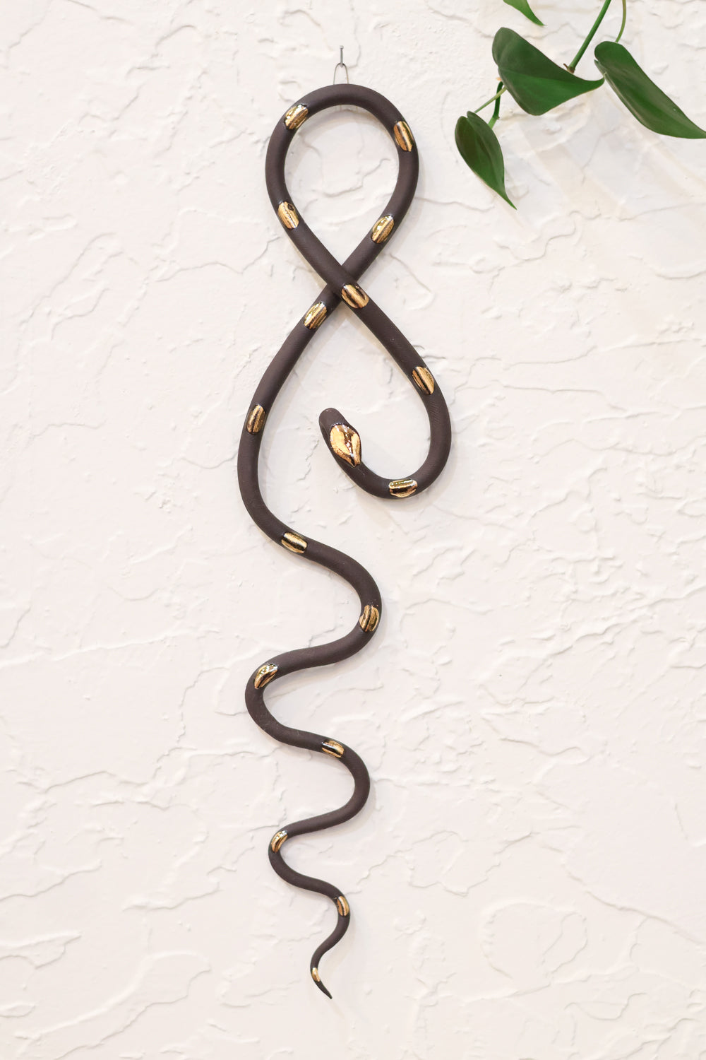 Black midnight clay vertical wall snake sculpture with gold glazed  head and dots. The top of the snake crosses like a figure 8 and the tail hangs down in an organic s curve. It hangs vertically from a small metal loop on a white wall with vining pothos. 