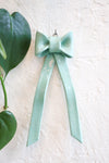 A glossy ceramic bow ornament in a pale mint green color, hanging on a textured off-white wall. A green heart-shaped leaf from a plant is visible to the left