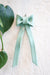 A hand holding a glossy ceramic ornament in the shape of a bow. The ornament is a deep blue with black edges, featuring a speckled texture. A leafy green plant is visible in the softly blurred background