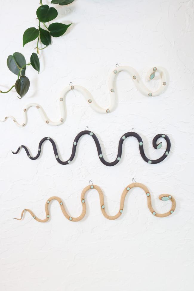 Three horizontal ceramic wavy snakes hanging on a white wall. The top snake is white clay with light turquoise glazed dots. The middle snake is a dark brown clay with light turquoise glazed dots. The bottom snake is tan clay with light turquoise glazed head and dots. 