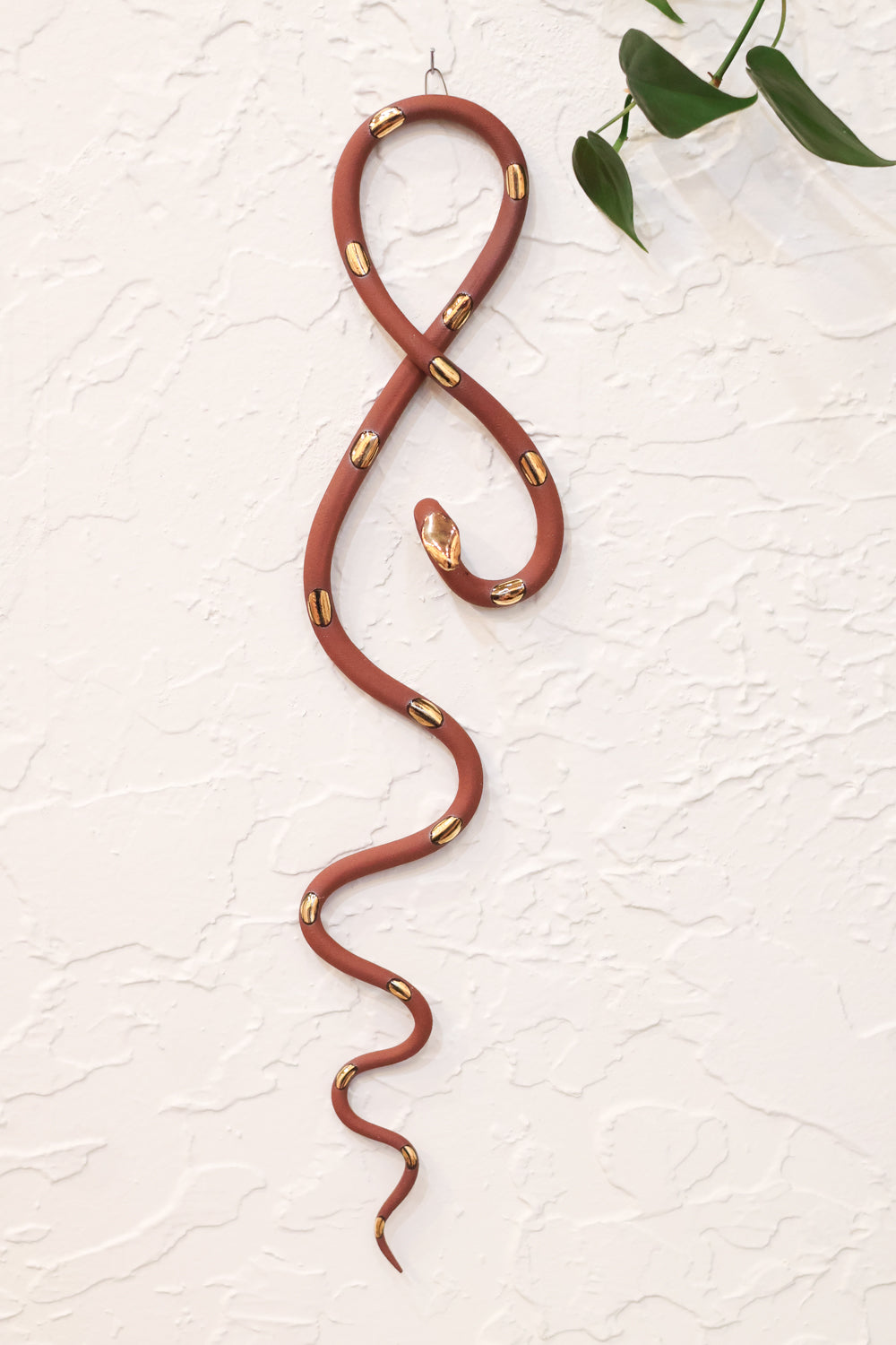 Red terracotta clay vertical wall snake sculpture with gold glazed  head and dots. The top of the snake crosses like a figure 8 and the tail hangs down in an organic s curve. It hangs vertically from a small metal loop on a white wall with vining pothos. 