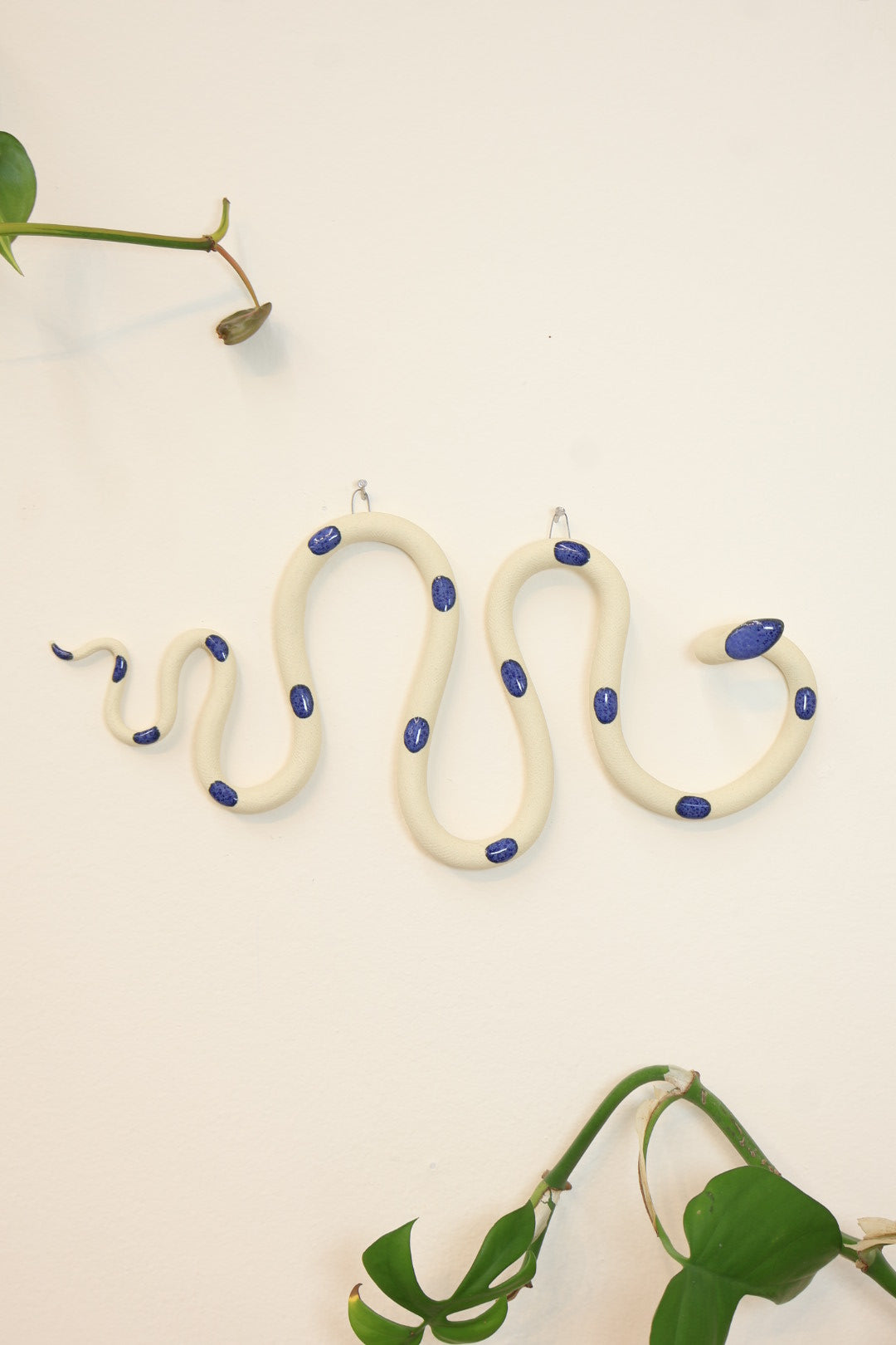 Horizontal wavy ceramic wall snake in the Selena colorway, ivory clay base and dark blue dots. It's hanging on a white wall with two small nails by its two small metal hooks.