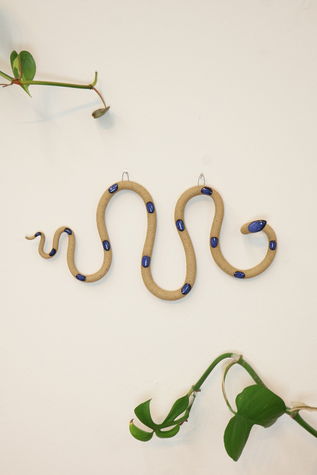 Horizontal wavy ceramic wall snake in the Selena colorway, speckled tan clay base and blue dots. It's hanging on a white wall with two small nails by its two small metal hooks.