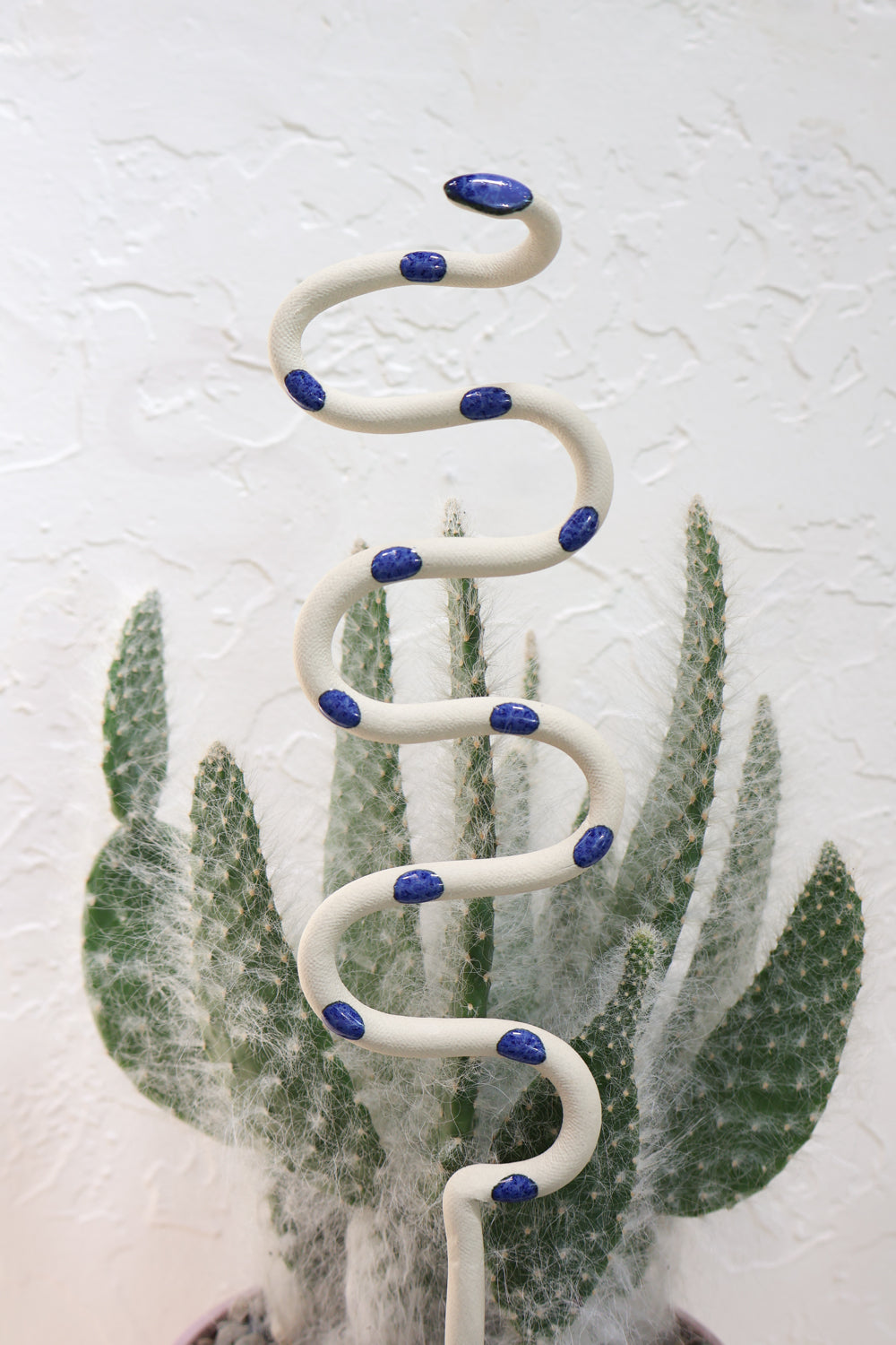 Three snake shaped plant stakes sticking out of a lavender pot with a cactus. There's a large black snake with green dots in the middle, and the medium sized snakes to each side. Tan with green spots snake to the left and a white snake with blue dots to the right. 