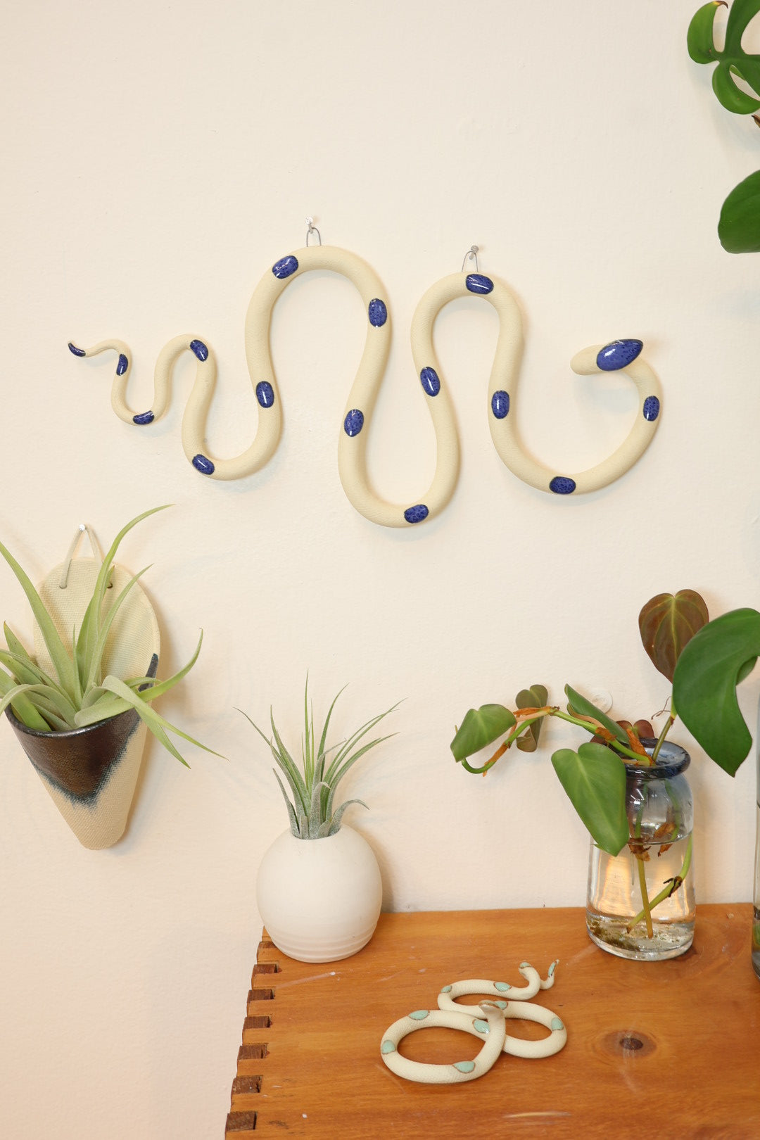 Styled horizontal Selena snake hanging on a wall next to an indigo wall planter. The snake hangs above a wooden shelf with glass propagation station with a plant, small white bud vase and a small Sylvia snake.