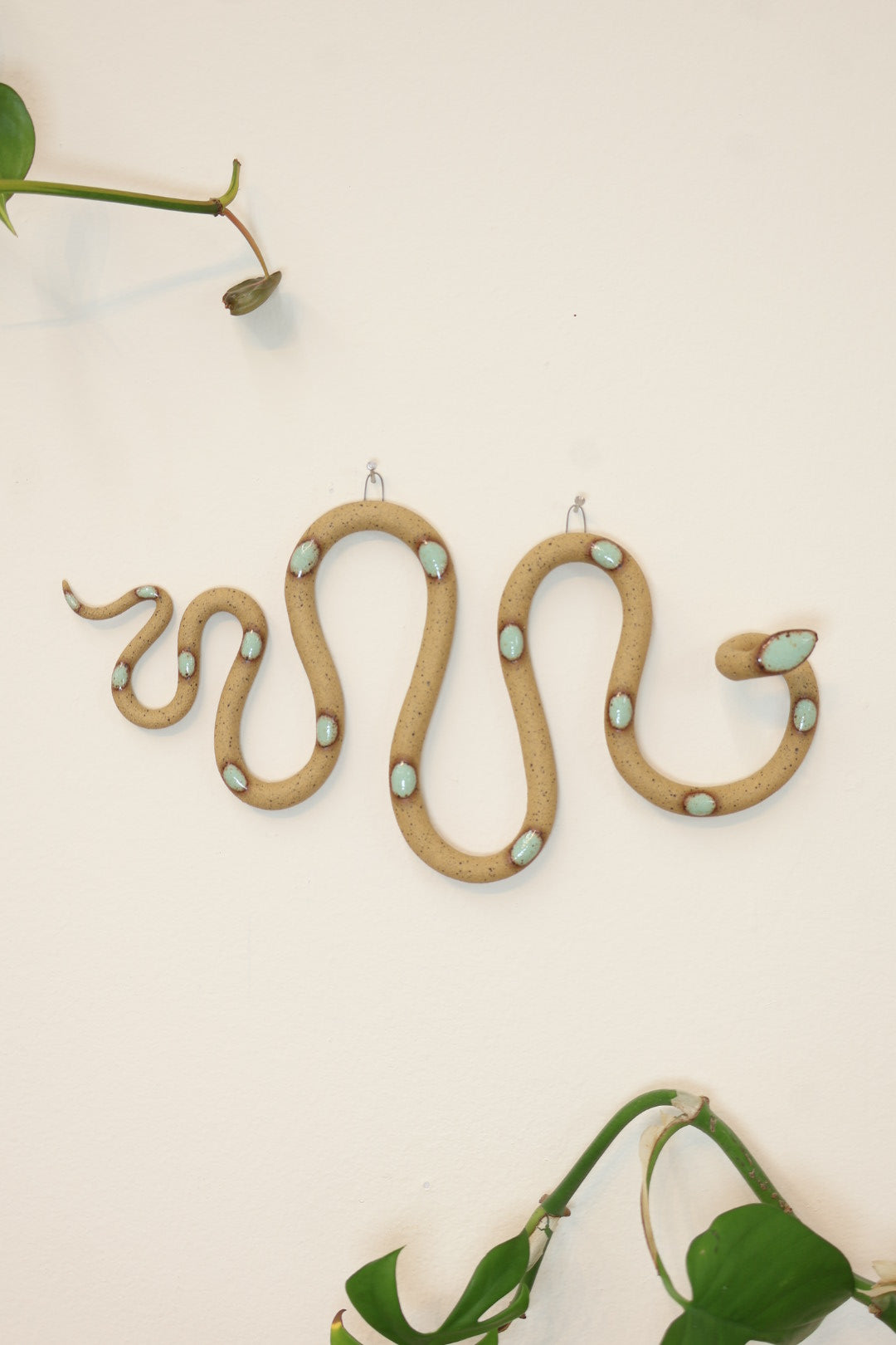 Horizontal wavy ceramic wall snake in the Selma colorway, speckled tan clay base and light turquoise dots. It's hanging on a white wall with two small nails by its two small metal hooks.