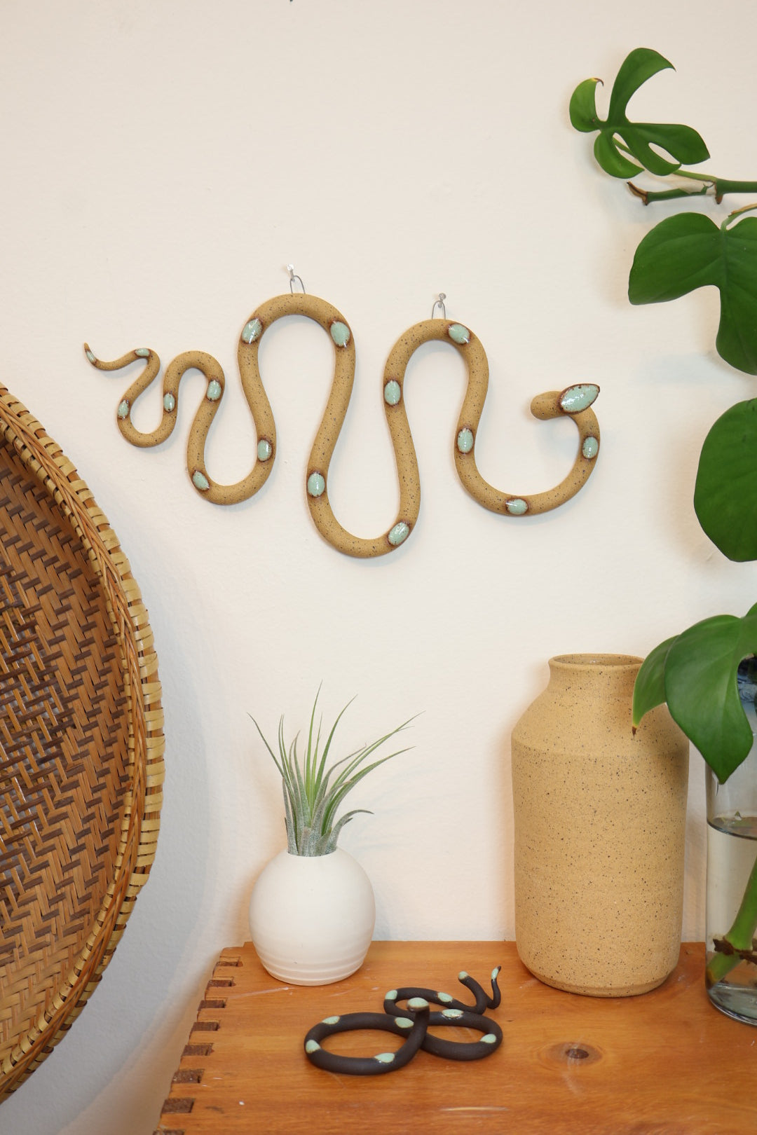 Decorative horizontal Selma snake hanging on a wall next to a woven basket. The snake hangs above a wooden shelf with a white bud vase, a speckled clay vase and a small Stevie snake. 