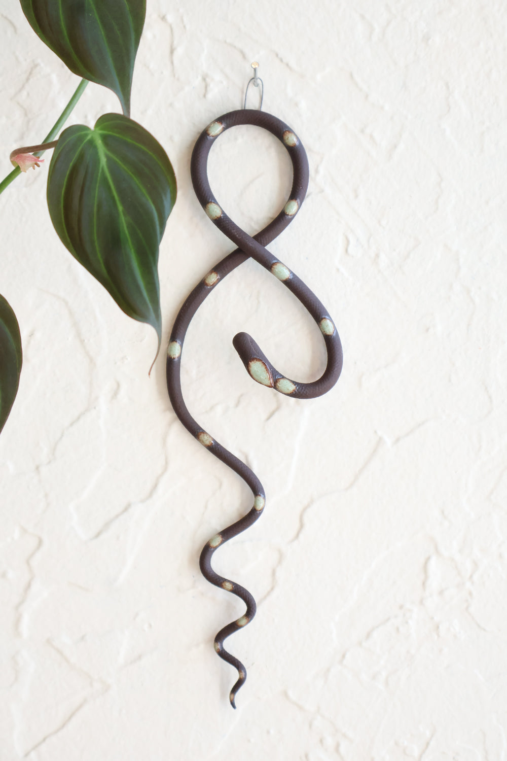 Small clay snake wall sculpture/decor hanging vertically from a metal loop on a white wall. The top of the snake crosses like a figure 8 and the tail hangs down in an organic s curve. This colorway is Stevie, dark brown clay and green turquoise dots. 