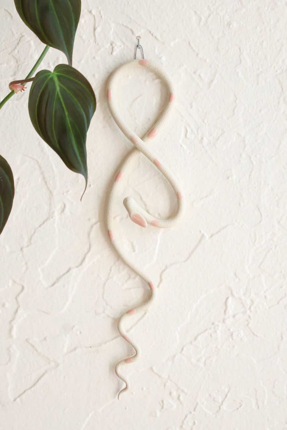 Small clay snake wall sculpture/decor hanging vertically from a metal loop on a white wall. The top of the snake crosses like a figure 8 and the tail hangs down in an organic s curve. This colorway is Sofie, ivory clay with light pink dots. 