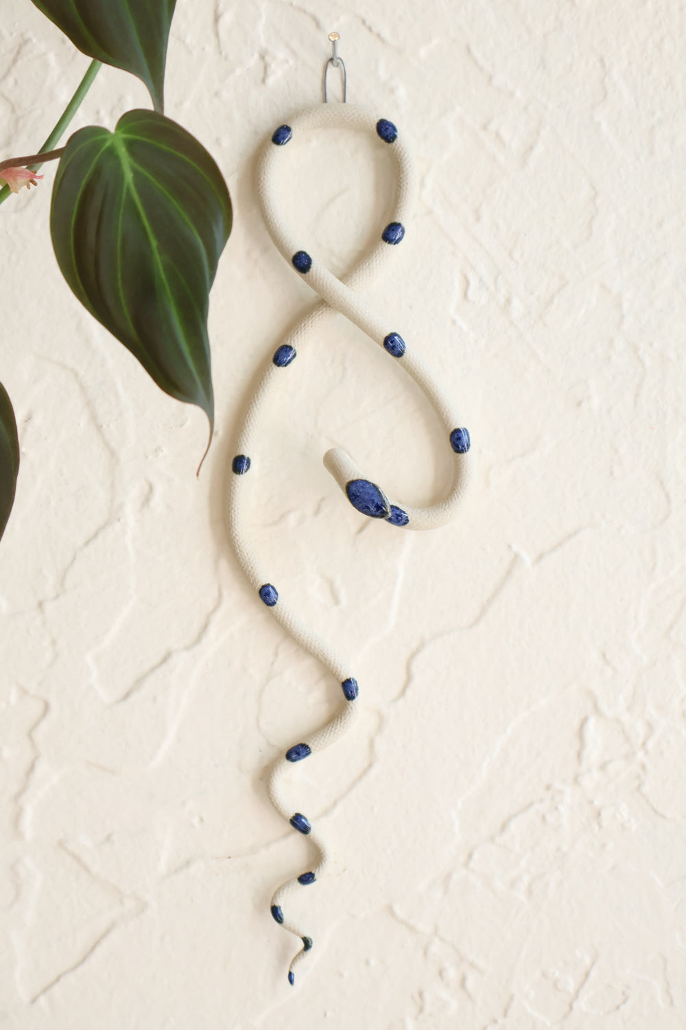 Small clay snake wall sculpture/decor hanging vertically from a metal loop on a white wall. The top of the snake crosses like a figure 8 and the tail hangs down in an organic s curve. This colorway is Selena, ivory clay with royal blue dots. 