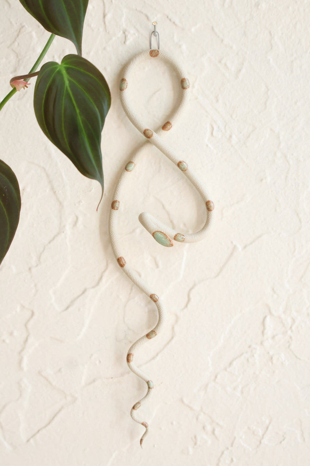 Small clay snake wall sculpture/decor hanging vertically from a metal loop on a white wall. The top of the snake crosses like a figure 8 and the tail hangs down in an organic s curve. This colorway is Sylvia, ivory clay with light green turquoise dots. 