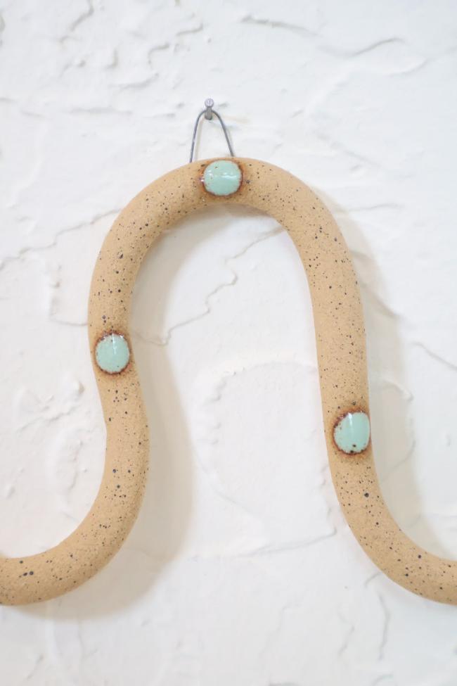 A close up of one of the ceramic snake curves showing the Selma colorway, speckled tan clay base and light turquoise dots. It hangs on a nail from a small metal loop.