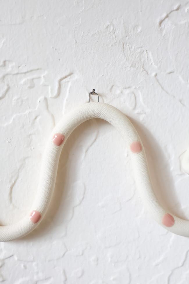A close up of one of the ceramic snake curves showing the Sofie colorway, ivory clay base and light pink dots. It hangs on a nail from a small metal loop.