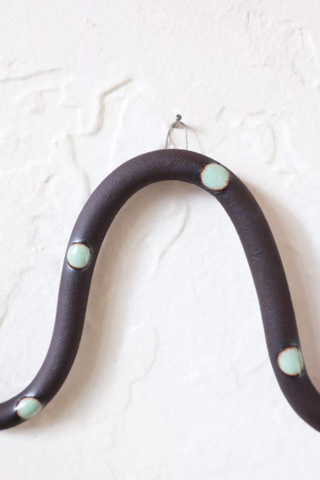 A close up of one of the ceramic snake curves showing the Stevie colorway, dark brown clay base and light turquoise dots. It hangs on a nail from a small metal loop.