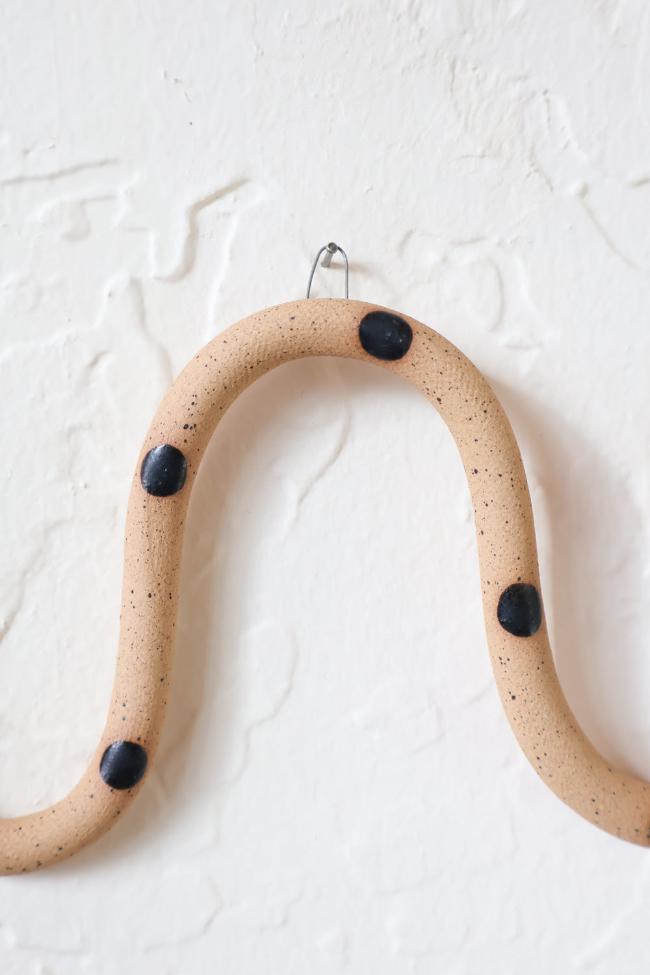 A close up of one of the ceramic snake curves showing the Sammy colorway, speckled tan clay base and black dots. It hangs on a nail from a small metal loop.