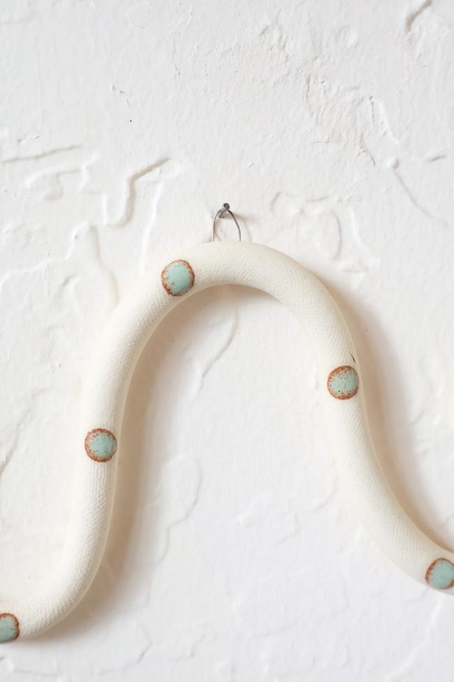 A close up of one of the ceramic snake curves showing the Sylvia colorway, ivory clay base and light turquoise dots. It hangs on a nail from a small metal loop.