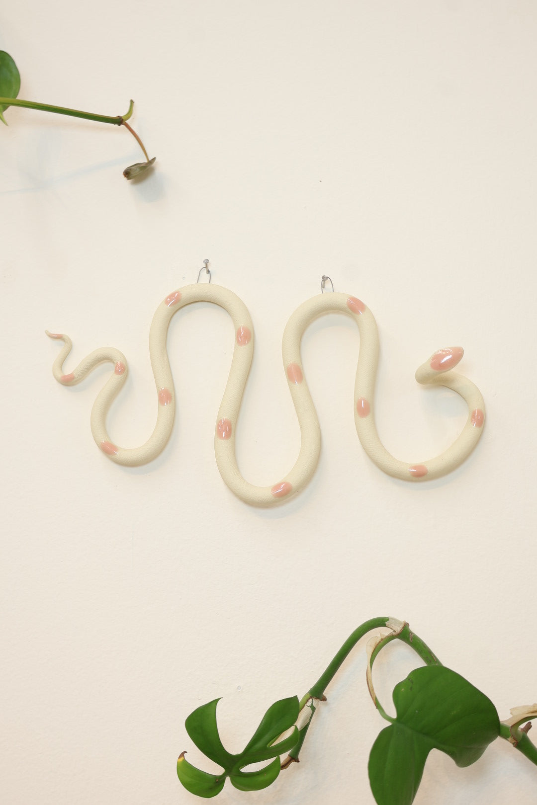 Horizontal wavy ceramic wall snake in the Sofie colorway, ivory clay base and light pink dots. It's hanging on a white wall with two small nails by its two small metal hooks.