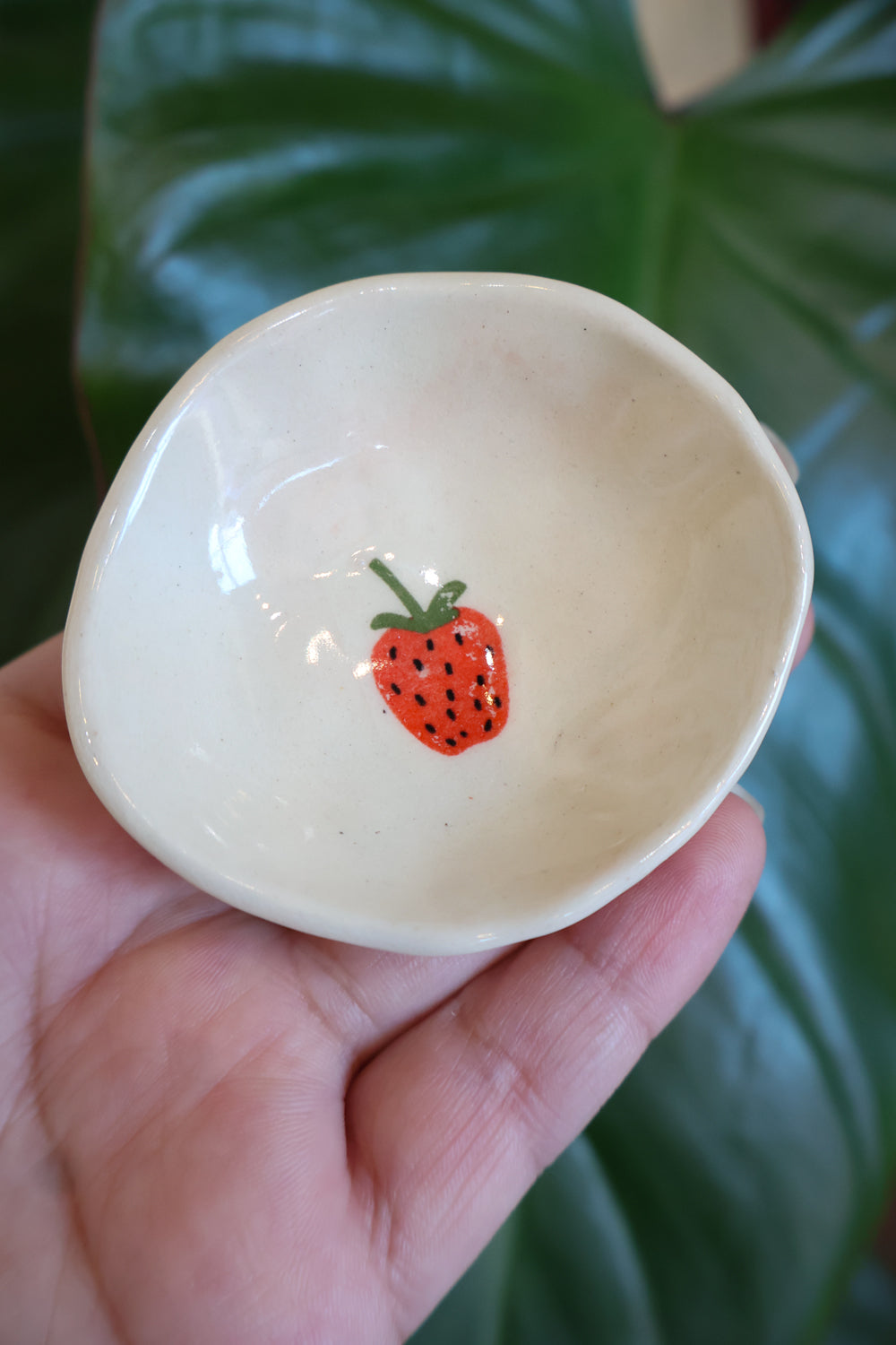 Small white dish with a pink strawberry detail. A red strawberry candle sits on the dish. There is a white background with green leaves.