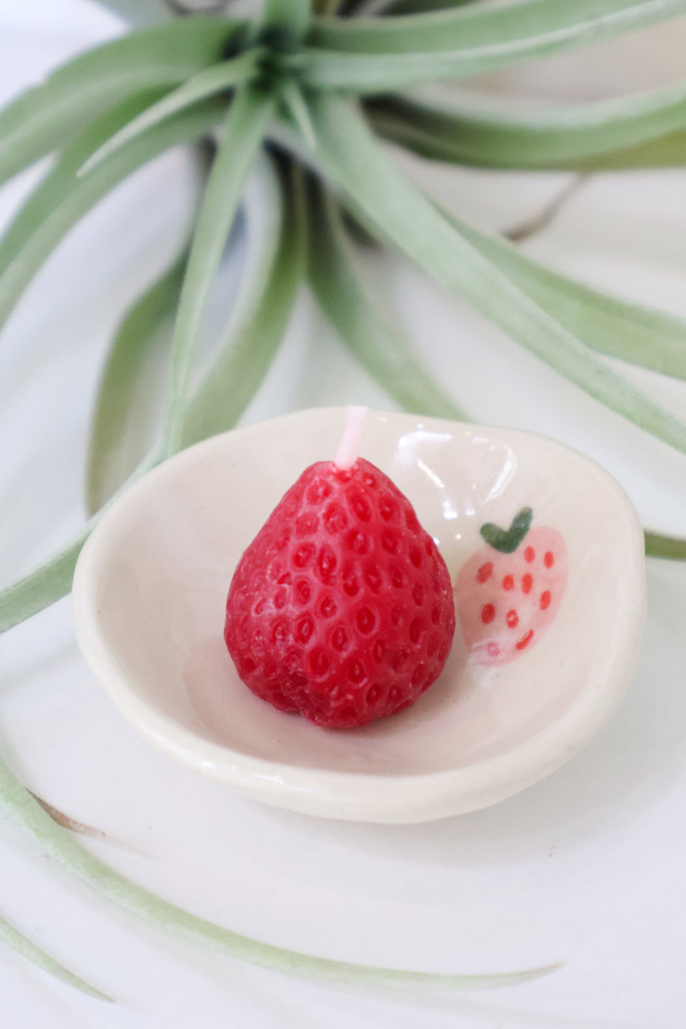 Small white dish with a pink strawberry detail. A red strawberry candle sits on the dish. There is a white background with green leaves.