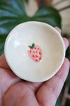Small white ceramic bowl with a single strawberry decal held in a hand against a blurred green background