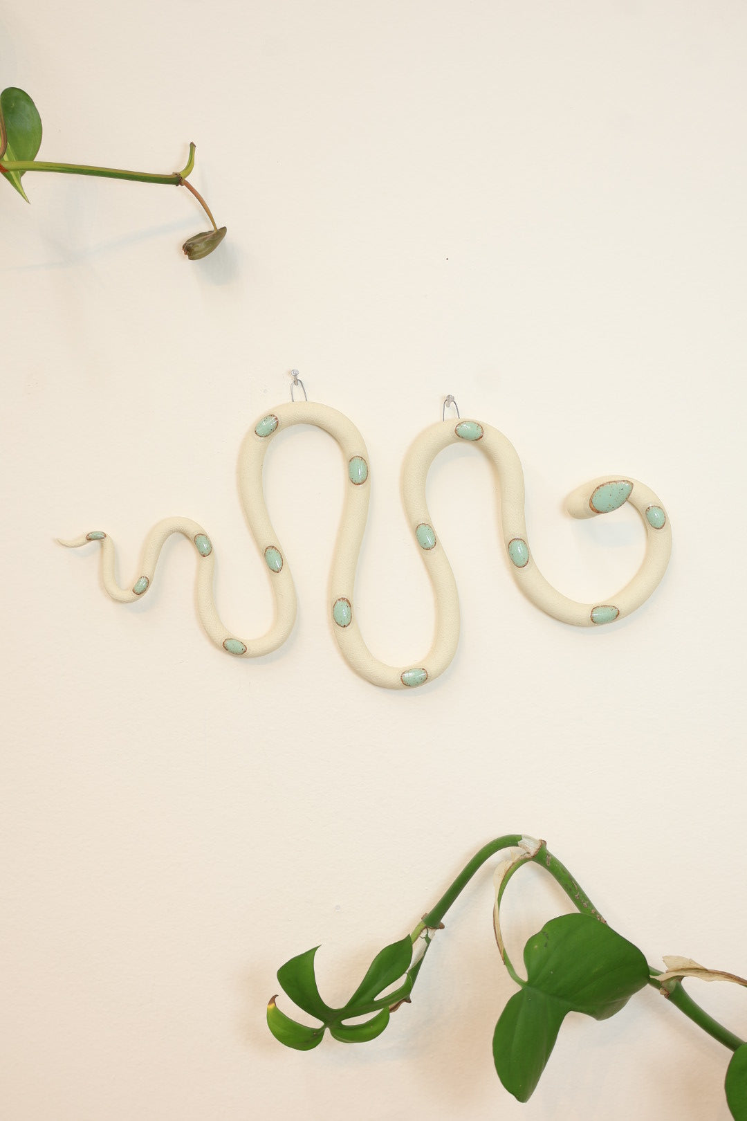 Horizontal wavy ceramic wall snake in the Sylvia colorway, ivory clay base and light turquoise dots. It's hanging on a white wall with two small nails by its two small metal hooks.