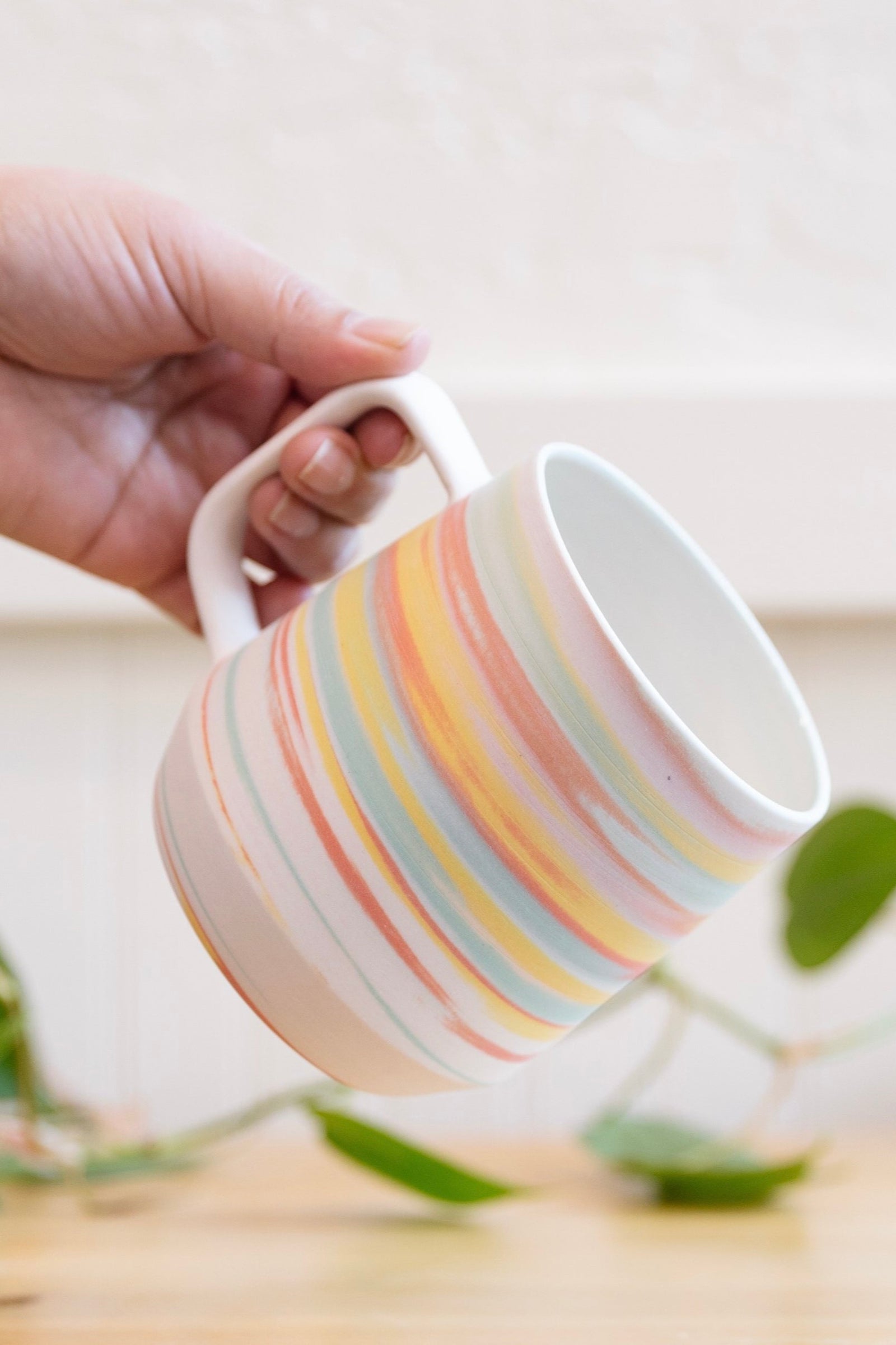 A hand holding a ceramic mug with pastel rainbow stripes in shades of yellow, pink, orange, and teal blue. The mug features a clean white handle, with a matte finish. The background includes a blurred green plant and a light-colored wooden surface