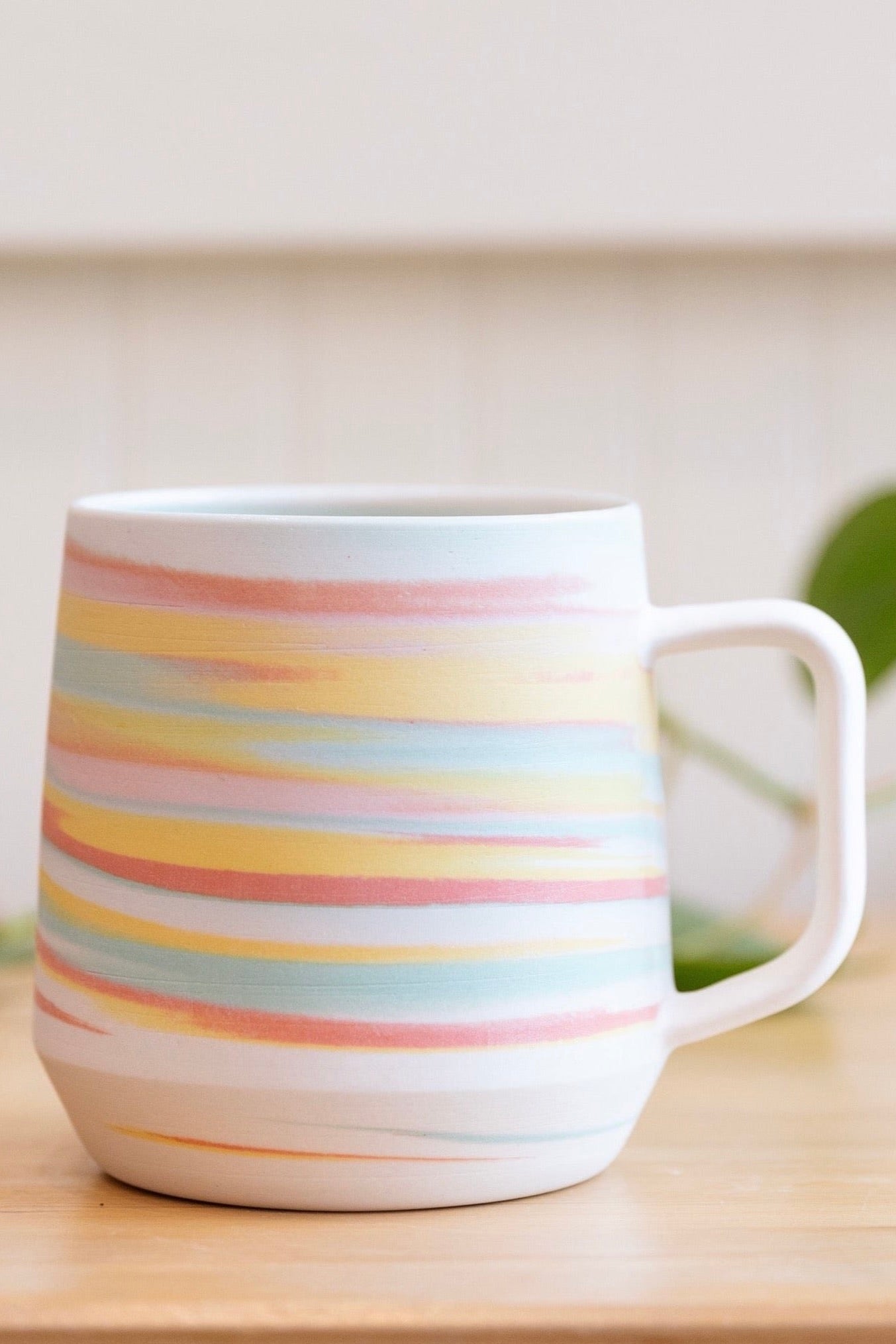 A hand holding a ceramic mug with pastel rainbow stripes in shades of yellow, pink, orange, and teal blue. The mug features a clean white handle, with a matte finish. The background includes a blurred green plant and a light-colored wooden surface