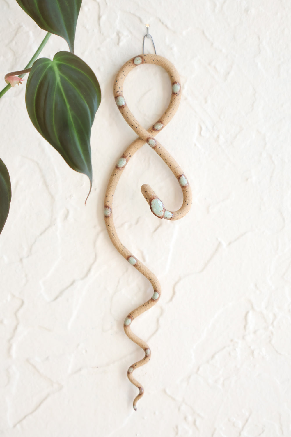 Small clay snake wall sculpture/decor hanging vertically from a metal loop on a white wall. The top of the snake crosses like a figure 8 and the tail hangs down in an organic s curve. This colorway is Selma, speckled tan clay with green turquoise dots. 