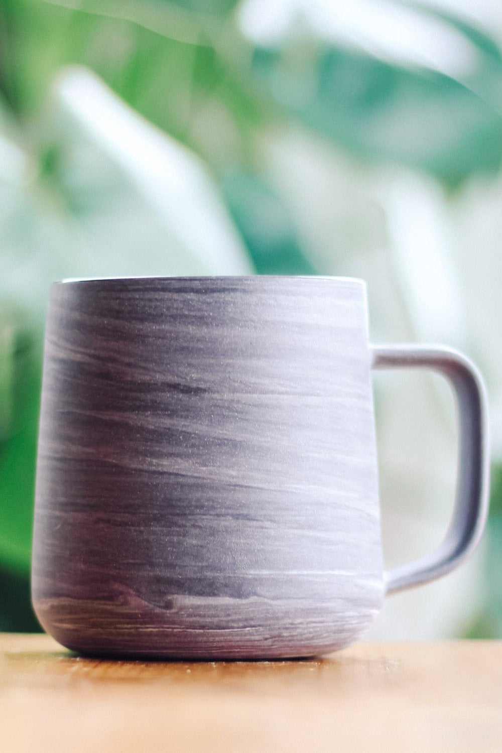 A modern grey ceramic mug with a matte finish featuring marbled stripes of white and black sits on a light wooden surface. The mug has a gently curved, cylindrical shape that tapers slightly towards the top and a sleek, straight handle.
The image background features soft, natural greenery with blurred large leaves.